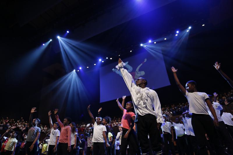 Many children singing with their right arm up on a blue-lit stage. An illustration of a man breakdancing is the backdrop.